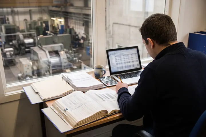 A controller works through printed ledgers and a spreadsheet at a desk overlooking the factory floor.
